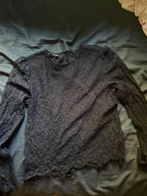 Svart spetsblus med lång ärm - Säljer en svart blus i spets med långärmad design och rund halsringning. Blusen har ett vackert blommigt spetsmönster och vågig kant nertill. Perfekt för dig som gillar en elegant och stilren look.