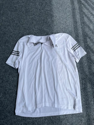 Vit pikétröja från Adidas - Snygg vit pikétröja från Adidas med klassiska grå ränder på ärmarna och liten Adidas-logga på bröstet. Tröjan har krage och korta ärmar, perfekt för sport eller träning. Tillverkad i ett lätt och ventilerande material.