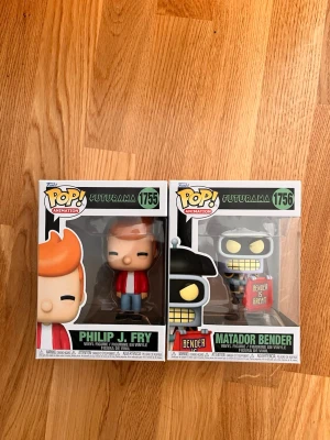 Funko Pop Futurama figurer - Två Funko Pop vinylfigurer från Futurama: Philip J. Fry (#1755) med röd jacka och blå byxor samt Matador Bender (#1756) med svart hatt och skylt. Båda figurerna är i sina originalkartonger och har tydliga detaljer från serien.