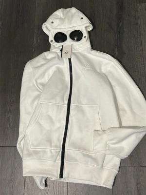 Vit hoodie med glasögon från C.P. Company - Säljer en vit hoodie från C.P. Company med hel dragkedja och ikoniska inbyggda glasögon i huvan. Hoodien har känguruficka framtill och ribbade muddar. Perfekt för dig som vill sticka ut med en unik streetwear-look.