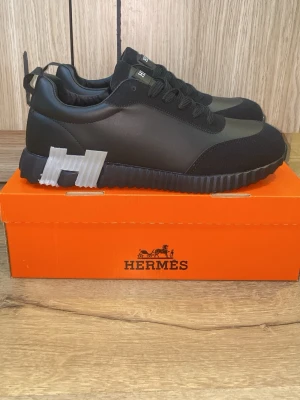 He*mès Bouncing sneaker - Snygga svarta sneakers från He*mès med stor vit H-detalj på sidan och sulan. Skorna har en låg profil, snörning och är tillverkade i en mix av läder och textil. Perfekta för dig som vill ha en clean och exklusiv look.