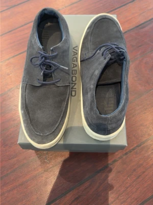 Mörkblå mocka loafers från Vagabond - Snygga vintage mörkblå loafers från Matinique i mjuk mocka med vita, tjocka gummisulor. Skorna har klassisk seglarsko-look med rundad tå och snörning framtill. Perfekta för en avslappnad men stilren outfit.