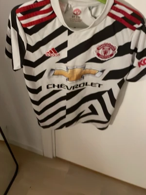 Manchester United zebra-matchtröja - Säljer en Manchester United matchtröja från Adidas med svartvitt zebramönster, röda detaljer och klubbmärke på bröstet. Tröjan har korta ärmar, rund hals och är tillverkad i ett lätt, ventilerande material. Perfekt för fotbollsträning eller att bära till vardags.