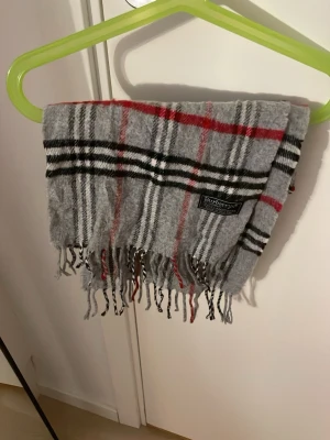 Grå rutig halsduk från Burberry - Klassisk grå halsduk från Burberry med rutigt mönster i svart, vitt och rött. Halsduken har fransar i ändarna och är tillverkad i mjukt material som känns lyxigt och stilrent. Perfekt accessoar för att lyfta din outfit.
