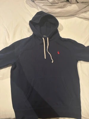 Mörkblå hoodie från Polo Ralph Lauren - Säljer en mörkblå hoodie från Polo Ralph Lauren med vit dragsko i huvan och klassisk röd logga broderad på bröstet. Hoodien har känguruficka framtill och är gjord i mjukt bomullsmaterial. Perfekt för en avslappnad och stilren look.