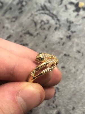 Guldfärgad ormring med diamant liknande stenar stämplad 18k och 750 - Cool ring i guldfärg formad som en orm, dekorerad med små glittrande stenar och gröna detaljer vid ögonen. Ringen har en slingrande design som ger ett unikt och trendigt intryck. Ringen är guldpläterad men är stämplad som 18karat därför säljer jag den billigare än äkta 18k.