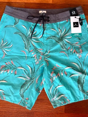 Badbyxor/Badshorts Nypris 599kr - Sprillans nya snygga turkosa badshorts från InterSport med tropiskt blommönster i grönt, rosa och vitt. Shortsen har grå midja med snörning och klassisk passform. Perfekta för stranden eller poolen, tillverkade i snabbtorkande material.
