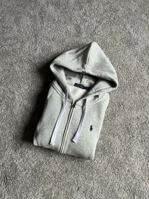 Grå zip hoodie från Polo Ralph Lauren - Snygg grå hoodie med dragkedja från Polo Ralph Lauren. Klassisk design med vit dragsko i huvan, två fickor fram och broderad logga på bröstet. Tillverkad i mjukt sweatshirtmaterial som är perfekt för chill dagar.