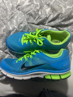 Nike skor - Snygga blå Nike sneakers med neongröna snören och innersula. Skorna har svart swoosh-logga, meshmaterial för bra andning och vit sula med gröna och svarta detaljer. Perfekta för dig som gillar sportig stil och vill sticka ut.