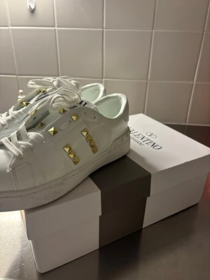 Vita Valentino Garavani sneakers - Snygga vita sneakers från Valentino Garavani med guldiga nitar på sidorna. Skorna är i bra skick har använt de ett få tals gånger ute annars bara testat inne. Priset kan diskuteras. Kontakta gärna om du är intresserad 