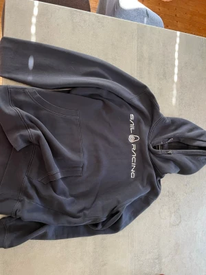  Sail Racing - Säljer en blå hoodie från Sail Racing med vit logga broderad på bröstet. Hoodien har klassisk känguruficka framtill och justerbar huva med snören. Perfekt för en avslappnad och sportig stil.