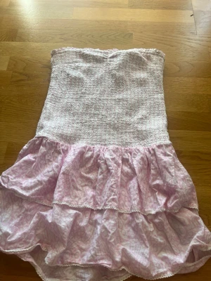 Ljusrosa axelbandslös kortklänning - Söt axelbandslös kortklänning i ljusrosa och vitt med volangkjol och smockad överdel. Klänningen har ett diskret mönster och är perfekt för varma dagar. Tillverkad i ett lätt och luftigt material som känns bekvämt mot huden. Endast använd fåtal gånger!🩷