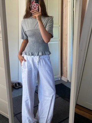Grå smockad topp med volangkant - Snygg grå topp med smockad midja och volangkant nedtill. Toppen har korta ärmar och rund halsringning. Materialet känns mjukt och stretchigt, perfekt för en chill och trendig look.