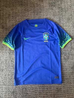 Blå Brasilien Nike fotbollströja VM 2022 - Snygg blå Brasilien fotbollströja från Nike med gröna och gula detaljer på ärmarna och leopardmönster. Tröjan har korta ärmar, rund hals och Brasilien-emblem på bröstet. Perfekt för fotbollsträning eller att bära som supporter. Lite stor i storlek passar M, Brasilien bortaställ 2022 VM
