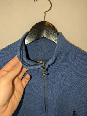 Blå stickad tröja med dragkedja Polo i pimabomull! - Snygg blå stickad tröja från Polo med halv dragkedja och hög krage. Tröjan har ett diskret broderat logotyp på bröstet och är tillverkad i ett mjukt, strukturerat material. Perfekt för lager på lager och enkel att matcha med jeans eller chinos. Enorm rabbat säljer för att jag flyttar allt ska bort, plagget är i original skick.