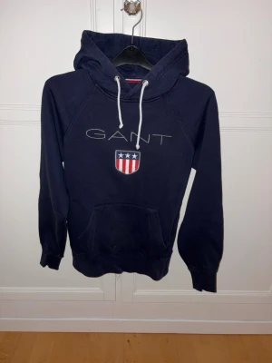 Mörkblå hoodie GANT - Mörkblå hoodie från GANT med vit dragsko i huvan och broderad logga samt sköld på bröstet. Klassisk känguruficka framtill och ribbade muddar vid ärmslut och nederkant. Perfekt för en avslappnad och sportig stil. Liten i storleken