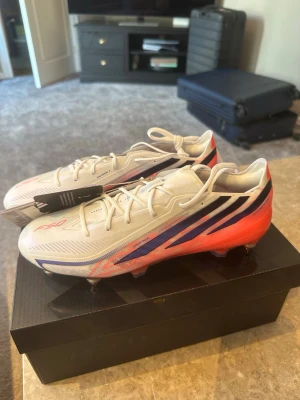Vita och röda fotbollsskor F50 - Snygga fotbollsskor i vitt med röda och blå detaljer. Skorna har låg profil, snörning och dobbar för bra grepp på planen, de är skruvdobbar och det är bäst att använda på gräs. Lätta material och sportig design med F50- och HYPERFAST-tryck på sidan. Perfekta för dig som vill ha snabbhet och stil på fotbollsplanen. De är också den nyaste modellen 