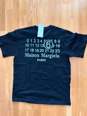 Maison Margiela t-shirt - Cool svart t-shirt från Maison Margiela med ikoniskt tryck på ryggen och diskreta vita stygn på bröstet. Klassisk rund hals och korta ärmar. Tillverkad i mjuk bomull som känns skön mot huden. Perfekt för dig som gillar streetwear och designerplagg.