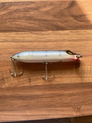 Två fiskedrag i olika färger - Säljer två fiskedrag med klassisk design. Ena draget är silverfärgat med blå rygg, röda detaljer och svart öga, medan det andra är mörkbrunt med gula ögon och guldfärgade prickar. Båda har två trekrokar i metall och är perfekta för fiskeentusiasten.