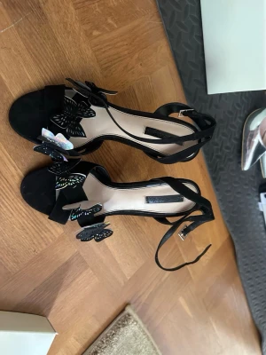 Svarta sandaler med fjärilsdetaljer - Unika svarta sandaler med höga klackar och flera dekorativa fjärilar i svart och holografiskt material. Skorna har smala remmar runt ankeln och öppen tå. 