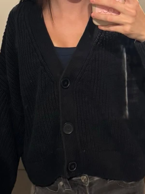 Oversized svart stickad kofta - Säljer en oversized svart stickad kofta med stora knappar framtill och djup V-ringning. Koftan har grovstickad struktur och långa ärmar, perfekt för lager på lager och en chill look.