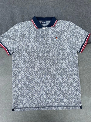 Blommig pikétröja från Jack & Jones - Snygg blå och vit pikétröja från Jack & Jones med blommigt mönster. Tröjan har klassisk krage med röd och vit rand, tre knappar framtill och korta ärmar med matchande detaljer. Perfekt för en trendig och avslappnad stil.