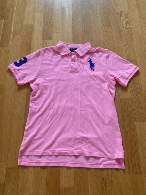 Rosa Polo Ralph Lauren pikétröja - Säljer en rosa pikétröja med klassisk krage och knappar framtill. Tröjan har korta ärmar, blå broderad logga på bröstet och siffran 3 på ena ärmen. Perfekt för en sportig och avslappnad stil.Polo Ralph Lauren loggan är lite nopprig som du kan se på bilden men den är inte så mycket nopprig.Sista bilden kan du se hur den passar på mig som är 177 cm lång.Storleken är typ mellan XS och S.Jag kan gå ner i pris