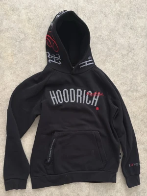 Svart hoodie från Hoodrich med tryck - Svart hoodie från Hoodrich med stor logga på bröstet och broderade detaljer i vitt och rött. Huva med tryck och känguruficka framtill. Texttryck även på ryggen och ärmen. Perfekt för en streetwear-look.