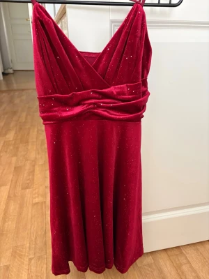 Röd glittrig klänning - Snygg röd klänning med omlottdesign och glittriga detaljer. Klänningen har en markerad midja och axelband, samt ett mjukt, sammetsliknande material som ger en lyxig känsla. Perfekt för dig som vill sticka ut med stil. Storlek S, aldrig använd😊.