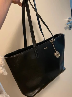 Svart shoppingväska från DKNY - Snygg svart shoppingväska från DKNY i slätt läder med dubbla axelband och guldfärgad logga samt hängande DKNY-berlock. Perfekt storlek för att få plats med allt du behöver och stilren design som passar till det mesta.
