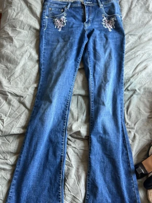 Blå bootcut jeans med broderi - Säljer ett par blå bootcut jeans med snygga broderade detaljer i vitt och rosa både fram och bak. Jeansen har klassisk femficksmodell och är tillverkade i denim. Perfekta för dig som gillar en retro och unik stil!