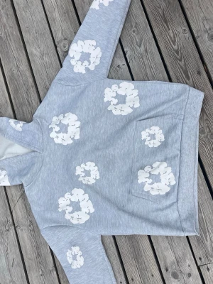 Ljusgrå hoodie med vita blommor - Säljer en ljusgrå hoodie med stora vita blommönster över hela tröjan. Hoodien har en klassisk huva och en stor magficka framtill. Mjuk och skön bomullsblandning, perfekt för en avslappnad stil. Passar dig som gillar unika prints och streetwear vibes.