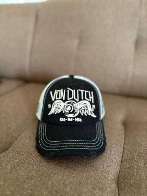 Svart och vit Von Dutch keps - Cool truckerkeps från Von Dutch i svart och vitt med broderad logga och vingmotiv framtill. Kepsen har vit mesh på sidorna och justerbar snapback baktill. Perfekt för en streetwear-look.