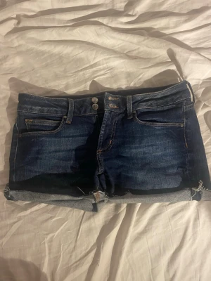 Mörkblå jeansshorts med uppvik - Säljer ett par mörkblå jeansshorts med uppvikta benslut och dubbla knappar i midjan. Shortsen har klassiska fem fickor och snygga kontrastsömmar baktill. Perfekta för sommaren och lätta att matcha med olika stilar.
