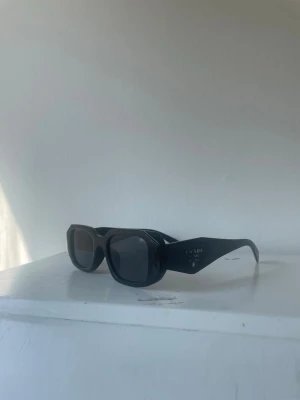 Prada solglasögon  - Snygga svarta solglasögon från Prada med rektangulära bågar och breda skalmar. Glasen är mörka och bågarna har en stilren, chunky design som ger en cool vibe. Perfekta för dig som vill ha en statement-accessoar till sommaren.