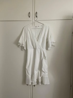 Vit omlottklänning med volang - Snygg vit kortklänning med omlottmodell och v-ringning. Klänningen har korta, vida ärmar och volangdetaljer längs nederkanten. Perfekt för sommarens alla tillfällen och ger en fräsch och luftig känsla.