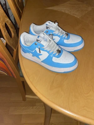 BAPE STA skor - Säljer ett par BAPE STA sneakers i blått och vitt med klassisk stjärndesign på sidan. Skorna har vita snören, perforerad tå och silverdetaljer på snörningen. Ovandelen är i läder och sulan är vit med blå detaljer. Perfekt för dig som gillar streetwear. Pris kan sänkas lite vid snabb affär. De är använda utomhus 1-2 gånger.