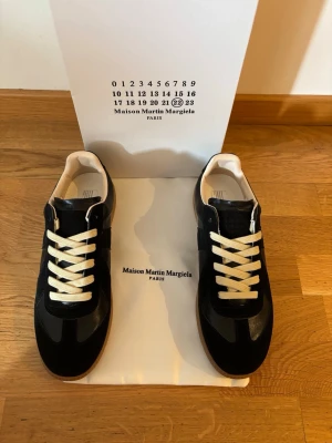 Maison Margiela svarta sneakers - Svarta sneakers från Maison Martin Margiela med vita snören och beige insida. Skorna har en klassisk låg siluett och är tillverkade i en mix av läder och mocka. Snygg och stilren design med diskreta detaljer och gummisula.