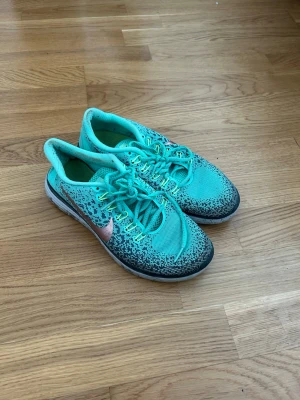 Turkosa Nike sneakers med rosa swoosh - Snygga turkosa sneakers från Nike med rosa swoosh på sidan. Skorna har mesh-ovandel för bra ventilation och svart mönsterdetalj runt tån och sidorna. Låga i modellen och med vita och svarta detaljer på sulan. Perfekta för en sportig look.