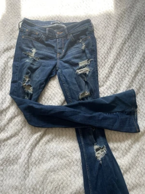 Mörkblå lågmidjade jeans med hål  - Säljer ett par mörkblå jeans med bootcut-modell och lågmidjade och flera hål på framsidan. Jeansen har klassisk femficksdesign och är gjorda i denim. Bootcut sytt själv. 