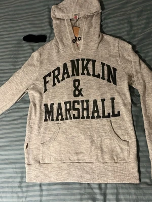 Grå hoodie från Franklin & Marshall - Snygg grå hoodie från Franklin & Marshall med stor svart logga framtill. Klassisk känguruficka och huva med dragsko. Tillverkad i mjukt sweatshirtmaterial, perfekt för en avslappnad stil.