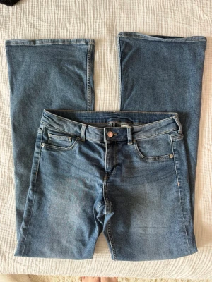 Blå bootcut jeans med fem fickor - Säljer ett par klassiska blå bootcut jeans med fem fickor och normalhög midja. Jeansen har en snygg tvätt med lätt slitning framtill och stängs med knapp och dragkedja. Perfekta för en avslappnad och trendig look.