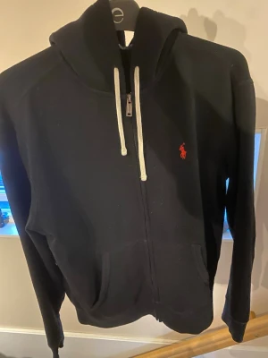 Svart ralph Lauren zip hoodie - Svart hoodie med dragkedja och vita snören, samt röd broderad Ralph lauren logga på bröstet. Tröjan är i gott skick och har mycket små tecken på användning.