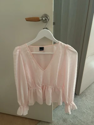 Ljusrosa blus med v-ringning - Söt ljusrosa blus med v-ringning och volang nedtill. Blusen har långärmade puffärmar med resår och små volanger vid ärmslut. Tillverkad i ett tunt, lätt och något transparent tyg🌷💞