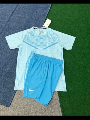 Ljusblått träningsset från Nike - Säljer ett ljusblått träningsset från Nike med t-shirt och shorts. T-shirten har ett mönster över bröstet och rund hals, medan shortsen är enfärgade med elastisk midja. Perfekt för träning eller sportaktiviteter.