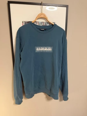 Blå sweatshirt från Napapijri - Säljer en blå sweatshirt från Napapijri med vit logga på bröstet. Tröjan har rund halsringning, långa ärmar och ribbade muddar vid ärmslut och nederkant. Perfekt för en avslappnad och stilren look.