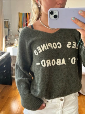 Mörkgrön stickad tröja med text - Mysig mörkgrön stickad tröja med vit text framtill som säger 'LES COPINES D'ABORD'. Tröjan har avslappnad passform, långa ärmar och bred halsringning som kan bäras off-shoulder. Perfekt för en chill och trendig look.