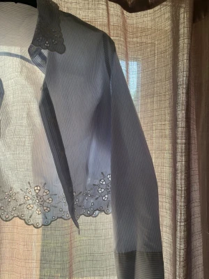Blåvit randig skjorta med spetsdetaljer - Säljer en blåvit randig skjorta med långa ärmar och fina spetsdetaljer på krage och nederkant. Skjortan har knappar framtill och ett elegant broderat blommönster som ger en unik touch. Perfekt för dig som gillar detaljer och stilren design.