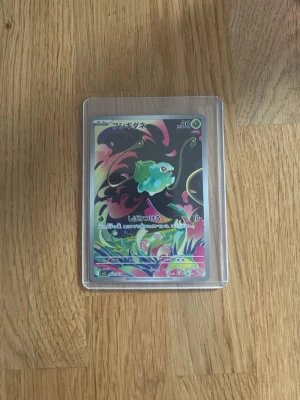 Japanskt Bulbasaur Pokémonkort - Unikt japanskt Pokémonkort med Bulbasaur-motiv. Kortet har färgstark design med svart, grön, rosa och gul bakgrund och ligger i ett genomskinligt plastfodral. Perfekt för samlare eller Pokémonfans som vill utöka sin samling med något speciellt.