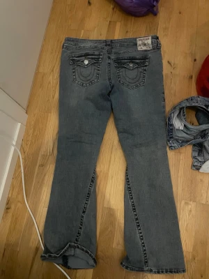 Grå bootcut jeans med fickdetaljer - Säljer ett par blå bootcut jeans med snygga fickdetaljer bak och kontrastsömmar. Jeansen har en klassisk femficksmodell och är tillverkade i denim med lätt tvättad look. Perfekta för dig som gillar en avslappnad men trendig stil.